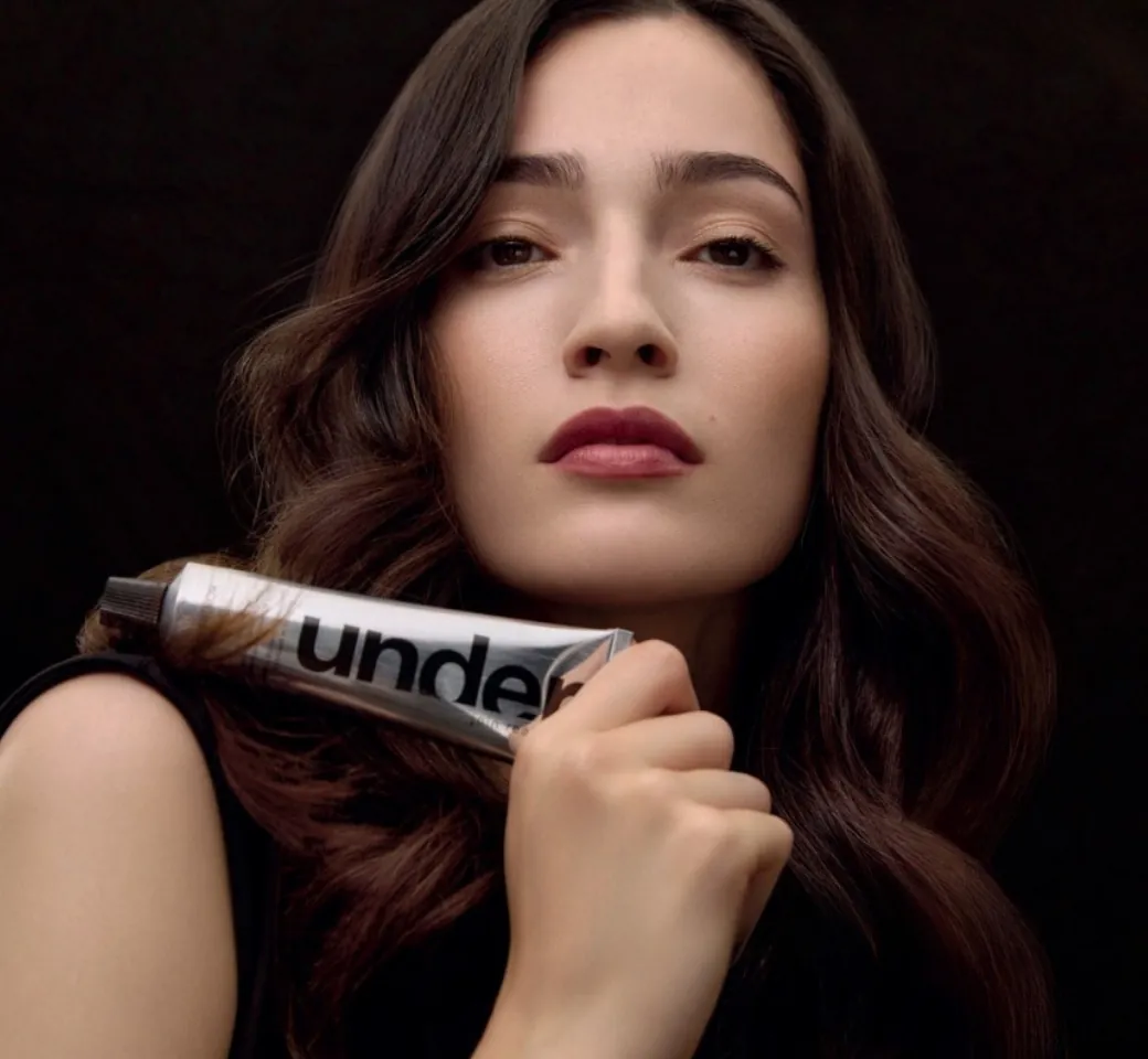 Model with long dark hair holding an Under colouring cream tube to her shoulder