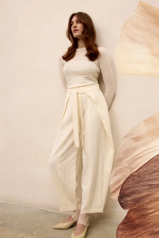 Model with long auburn hair in cream, standing against a wall beside a dried petal
